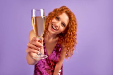 Parlak elbiseli, kıvırcık saçlı ve bir bardak şampanyası olan sevimli kızıl saçlı kadın mor arka planda izole bir şekilde eğleniyor. Parti, kutlama, doğum günü konsepti..
