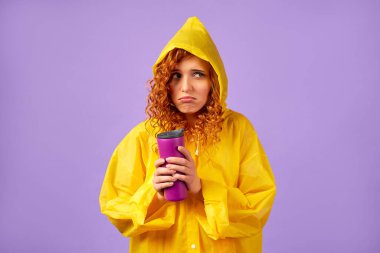 Sarı yağmurluklu kızıl saçlı kadın ve mor arka planda izole edilmiş termal bardak..