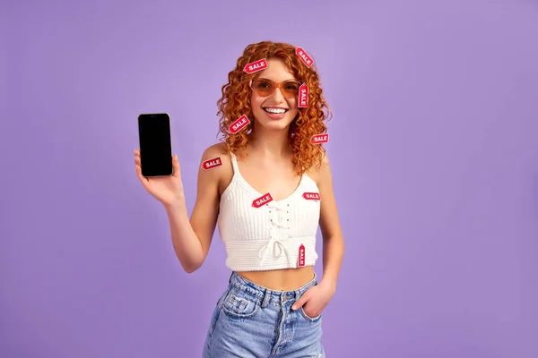 紫色の背景に隔離された空白のスマートフォン画面を示すカールと販売ラベルを持つかわいい赤い髪の女の子 ショッピングのコンセプト ショッピング — ストック写真