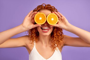 Kıvırcık saçlı tatlı bir kız mor arka planda izole edilmiş gözler yerine portakal yarımları tutuyor. Düzgün beslenme. Sağlıklı yaşam tarzı.