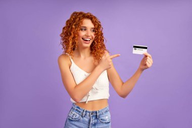 Kıvırcık saçlı şirin bir kız kredi kartı tutuyor ve onu işaret ediyor. Mor bir arka planda izole edilmiş..