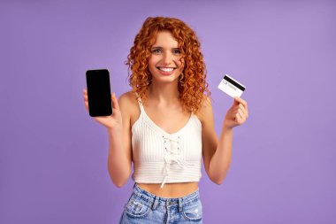 Kıvırcık saçlı tatlı bir kız kredi kartı tutuyor ve mor arka planda izole edilmiş siyah bir akıllı telefon ekranı gösteriyor. Çevrimiçi ödeme, bankacılık.
