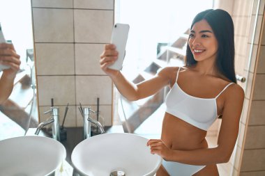 Beyaz iç çamaşırlı Koreli bir kadın banyoda aynanın önünde dikiliyor ve selfie çekiyor. Yüz ve vücut bakımı. Sabah rutini.