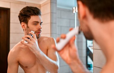 Beyaz havluyla güzel kaslı bir adam banyoda aynanın önünde duruyor ve yüzünü tıraş ediyor. Yüzüne ve vücuduna özen göster..