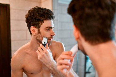 Beyaz havluyla güzel kaslı bir adam banyoda aynanın önünde duruyor ve yüzünü tıraş ediyor. Yüzüne ve vücuduna özen göster..