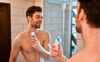 Beyaz havluyla güzel kaslı bir adam banyoda aynanın önünde duruyor ve yüzünü tıraş ediyor. Yüzüne ve vücuduna özen göster..