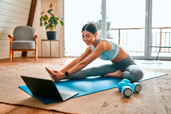 A sportive woman in sportswear does exercises, dumbbells lie nearby and uses a laptop at home in the living room. Sports and recreation concept.