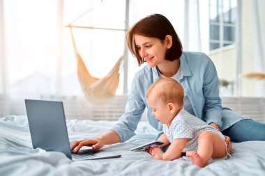 Evde kal, annesi uzaktan bilgisayarla çalışıyor, bebeğine bakıyor. Doğum izninde olan genç bir anne, küçük bir çocukla yatakta çalışmaya çalışır. Kapat, kopyala boşluk, arkaplan.