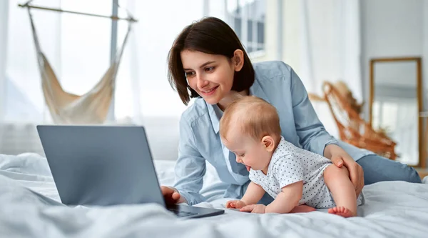 Stay at home, mom works remotely on laptop, taking care of her baby. A young mother on maternity leave tries to work in bed with a small child. Close up, copy space, background.