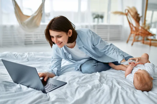 Stay at home, mom works remotely on laptop, taking care of her baby. A young mother on maternity leave tries to work in bed with a small child. Close up, copy space, background.