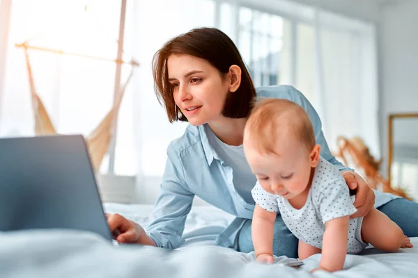 Stay at home, mom works remotely on laptop, taking care of her baby. A young mother on maternity leave tries to work in bed with a small child. Close up, copy space, background.