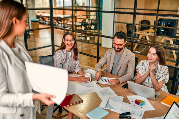 Focused young Caucasian business woman showing graph on paper to group of colleagues sitting at table with laptops in modern office.