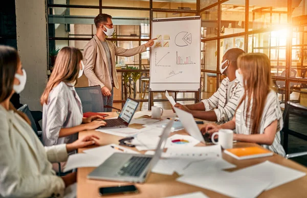 Workers discuss and meet while wearing a medical mask to protect against the corona virus. A young man writes a mind map on a whiteboard and makes new business plans during the COVID-19 pandemic.