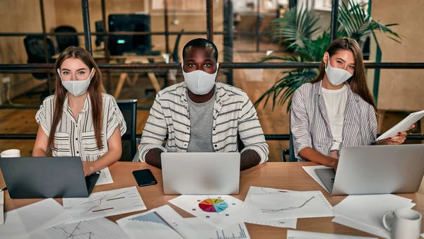 Office work in a pandemic and quarantine. A young group of multi-ethnic business people in protective masks are working on laptops in a modern office.