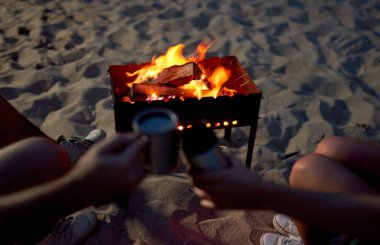 Ateşin yanında kupa bardak çay tutan bir adam ve kadının kesilmiş görüntüsü, deniz kıyısındaki kumlu bir sahilde..