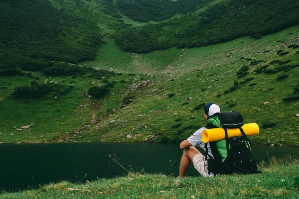 湖のそばの芝生にはリュックとゴム製のマットを持った男ハイカーが腰を下ろしている 緑の山々のふもとに若い観光客 — ストック写真