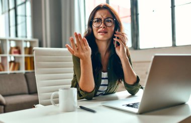 İş kadını evde bir masada oturup telefonla konuşurken dizüstü bilgisayarda çalışıyor. Serbest çalışmak, evden çalışmak..