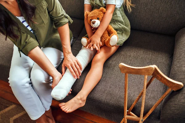 一个可爱的小女孩 腿断了 和妈妈坐在沙发上 抱着一只泰迪熊在家里 沙发附近有拐杖 可以快速康复 — 图库照片