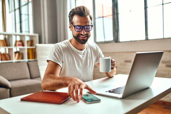 Businessman in glasses with a mug of coffee works at a laptop. Freelance, work from home.