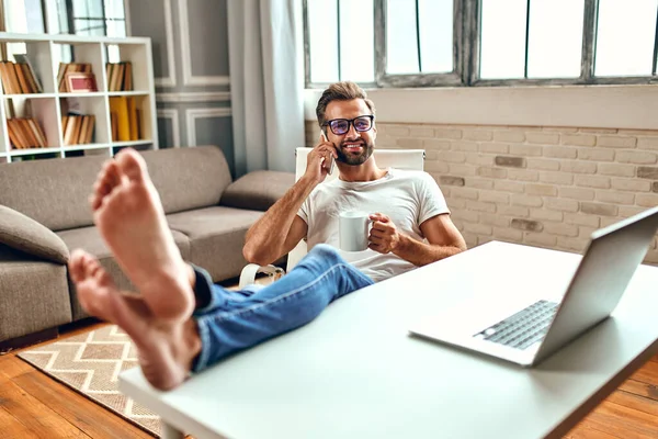 Businessman in glasses with a mug of coffee works at a laptop while sitting with his legs on the table. Freelance, work from home.