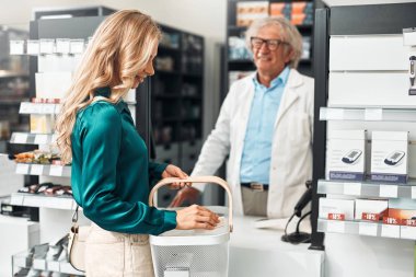 Beyaz önlüklü yaşlı bir erkek eczacı modern bir eczane tezgahında bayan bir müşteriye yardım ediyor. Gülümsüyorlar ve konuşuyorlar. Konsept: Müşteri hizmetleri, eczane, sağlık hizmetleri.