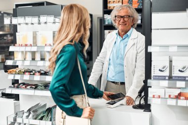 Gülümseyen kıdemli bir eczacı, eczane tezgahında akıllı telefon ödemesi yapan bayan bir müşteriye yardım ediyor. Konsept: erişimsiz ödeme, müşteri hizmetleri, sağlık hizmetleri, teknoloji.