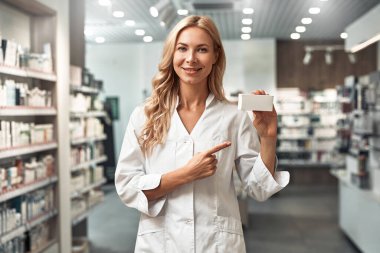 Beyaz üniformalı, güler yüzlü kadın eczacı modern bir eczane içinde dururken elinde boş bir ilaç kutusu tutuyor. Sağlık hizmetleri uzmanı ilaç sunumu konsepti.