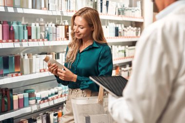 Uzun sarı saçlı genç bir kadın, tabletli bir profesyonelin yardımıyla modern bir eczanede alışveriş yaparken kozmetik şişesini tutuyor ve inceliyor..