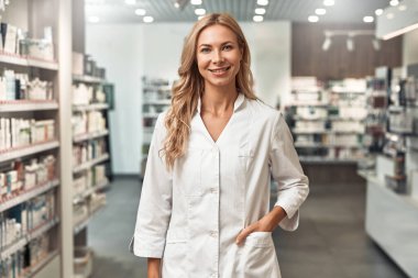 Beyaz önlüklü sarışın kadın eczacı modern bir eczanede duruyor, kendine güvenerek gülümsüyor. Profesyonel sağlık hizmeti kavramı.