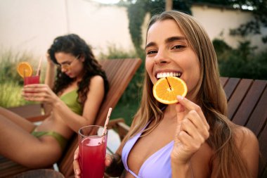 Arkadaşı dinlenme odasında dinlenirken, lavanta bikinili neşeli bir kadın portakal dilimini neşeyle ısırıyor ve meyve suyu tutuyor. Konsept: Tropikal eğlence, yaz tazeliği, gençlik.