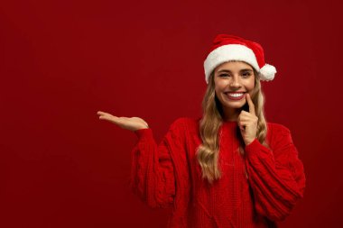 Kırmızı kazaklı ve Noel Baba şapkalı neşeli kadın kırmızı arka plandaki fotokopi alanını işaret ediyor. Şenlikli Noel portresi. Mutlu yıllar ve mutlu Noeller..
