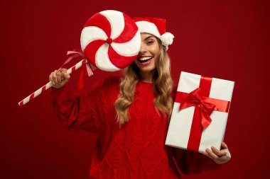 Noel Baba şapkası takan neşeli bir kadın, elinde hediye paketi tutan kırmızı bir kazak ve kırmızı arka planda naneli lolipop tutan bir kadın. Mutlu yıllar ve mutlu Noeller. Şenlikli kış tatili havası.
