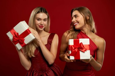 Kırmızı stüdyo arka planında kırmızı kurdeleli beyaz hediye kutuları taşıyan zarif kırmızı elbiseli iki neşeli kadın. Mutlu yıllar ve mutlu Noeller..