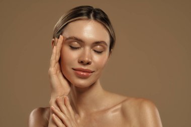 Yüzüne dokunan genç bir kadının yakın plan portresi. Çıplak makyaj, parlayan ten, doğal güzellik konsepti. Sıcak ışıklandırma ve nötr arkaplan cilt bakımını ve estetiği vurguluyor.