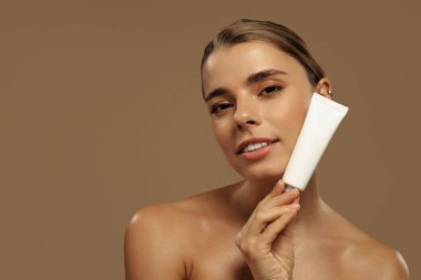 Yüzünde beyaz bir kozmetik tüp tutan parlak ciltli çekici genç bir kadın. Cilt bakımı, güzellik ürünü reklamı veya marka konsepti için ideal.