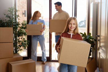 Gülümseyen ebeveynler ve kız yeni bir eve taşınırken karton kutular taşıyorlar. Parlak modern iç mekan, doğal gün ışığı. Taşınma kavramı, yeni başlangıçlar, aile takım çalışması..
