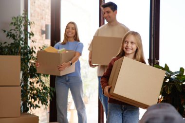 Gülümseyen ebeveynler ve kız yeni bir eve taşınırken karton kutular taşıyorlar. Parlak modern iç mekan, doğal gün ışığı. Taşınma kavramı, yeni başlangıçlar, aile takım çalışması..