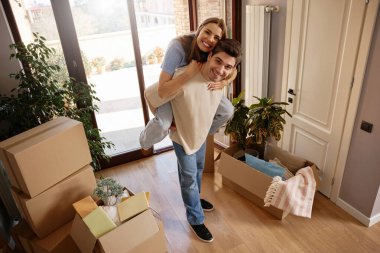 Hoş bir dairede, paketsiz kutuların arasında ortağının sırtına binen gülümseyen genç bir kadın. Çift taşınırken eğlenceli ve neşeli bir anın tadını çıkarıyor. Kavram: aşk, taşınma günü, ev, mutluluk.