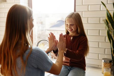 Anne ve kızı mutfak içinde el çırpma oyunu oynuyorlar. Sabah ışığı, kaynaşma ve sıcak aile ortamı..