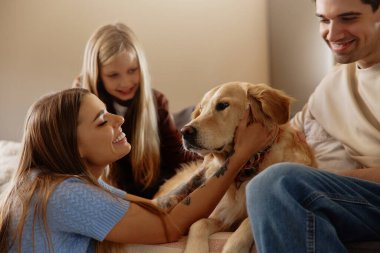 Genç bir kadın kanepede bir golden retriever 'a şefkatle sarılırken ailesi ona sıcakkanlılıkla ve neşeyle bakıyor. Kapalı mekanda kaynaşma anı. Konsept: evcil hayvanlar ve aile bağları için sevgi.