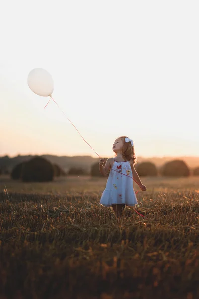 Dziewczynka w polu trzymając balon w świetle zachodu słońca w okresie letnim — Zdjęcie stockowe