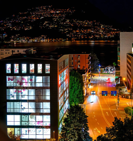 Ночной вид на Парадизо паромной станции и через озеро Лугано в Монте-Бре с подсветкой офисного здания Banca Credinvest Sa на переднем плане в городе Лугано в кантоне Ticio, Швейцария