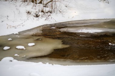 Buz, kar ve buzun altındaki kahverengi suyla ormandaki donmuş nehir manzarası. Kırsal kesimde kış yağmuru