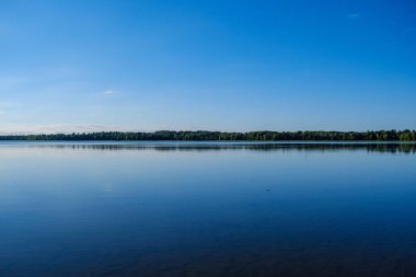 Geniş nehir manzarası geniş su kütlesi, gökyüzünün mavi yansımaları ve arka planda yeşil ağaçlar.