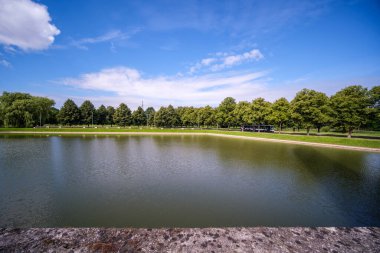 Geniş nehir manzarası geniş su kütlesi, gökyüzünün mavi yansımaları ve arka planda yeşil ağaçlar.