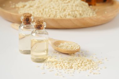 Küçük şeffaf şişelerde susam yağı ve tahta kaşığa dökülen susam tohumları beyaz arka planda..