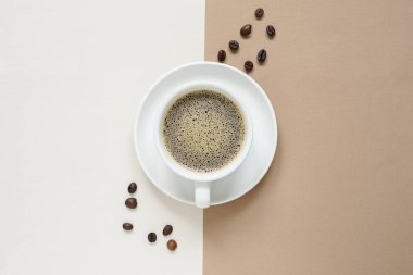 Sütlü kahve ve kahve köpüğünün üst görüntüsü beyaz seramik bir kahve bardağında ortadaki kavrulmuş kahve tepeciğinin yanına krema ve kahverengi arka plan arasına yerleştirilmiş beyaz seramik bardak..