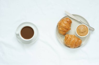 Beyaz seramik bardaktaki sıcak kahve fincanının üst görüntüsü ve beyaz seramik bardaklara yerleştirilmiş beyaz kruvasan ve fıstık ezmesi yanında beyaz kumaş, yuvarlak seramikte paslanmaz çelik bıçak.,.