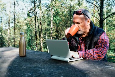 Olgun bir adam bardaktan içiyor, sıradan bir şekilde giyinip sahada dizüstü bilgisayarda çalışıyor.