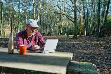 Olgun bir adam oturmuş, sıradan giyinmiş ve sahada bilgisayarla çalışıyor.